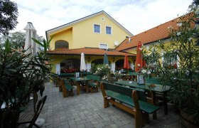 Gastgarten mit gr&uuml;nen Tischen und B&auml;nken sowie geschlossenen Sonnenschirmen vor einem gelben Geb&auml;ude.