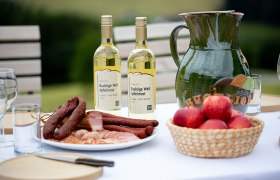 Tisch mit Apfelmost, Wurstplatte, Äpfeln, Holzbrett, Messer und Gläsern.