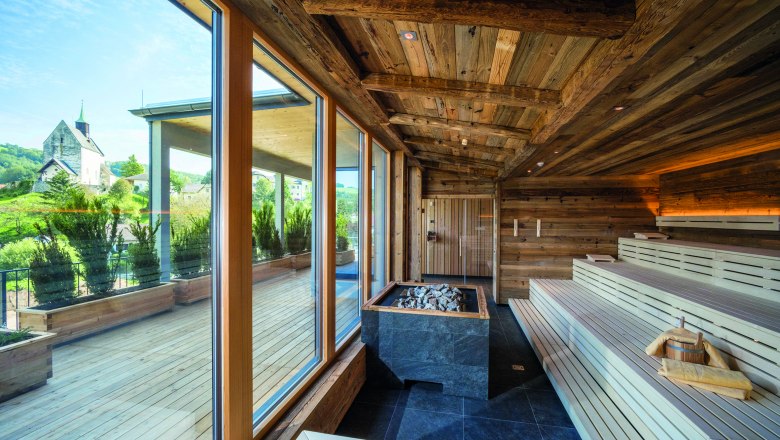 Innenansicht einer Sauna mit Holzb&auml;nken und gro&szlig;en Fenstern, die auf eine Terrasse und eine Kirche im Hintergrund blicken.