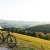 Ein Mann in weißem Hemd steht mit einem Fahrrad auf einem Hügel mit Blick auf eine grüne Landschaft.