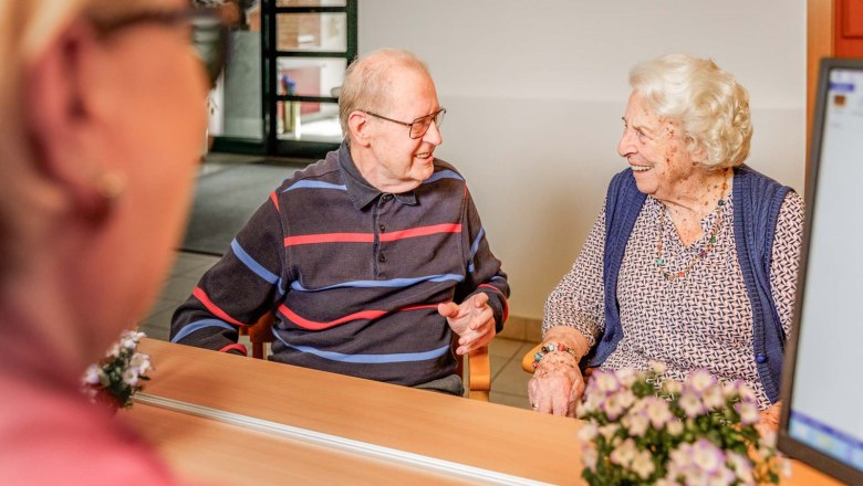 Two elderly people are sitting at a table smiling and talking in a cozy room.