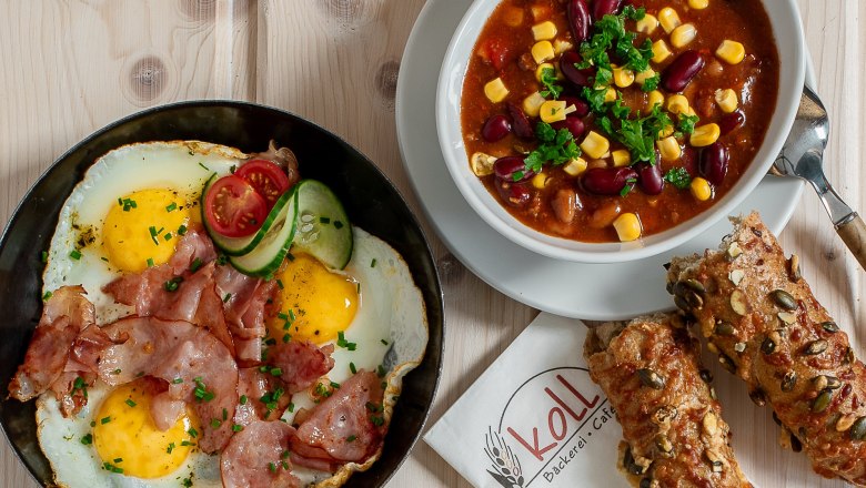 Verschiedene warme Snacks auf einem Tisch, darunter Spiegeleier mit Speck, Chili con Carne, belegte Brote und Brötchen.