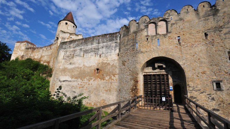 Eingangstor einer alten Burg mit Holztor und Zinnenmauer, blauer Himmel im Hintergrund.