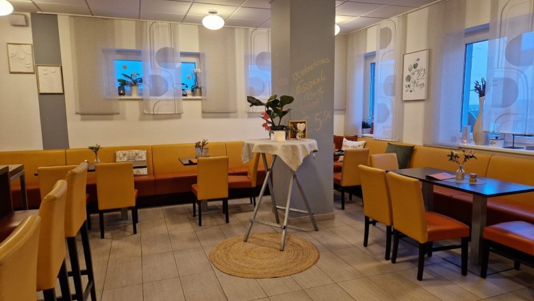 Innenansicht eines gemütlichen Cafés mit gelben Stühlen und Tischen, dekoriert mit Pflanzen und Bildern an den Wänden.