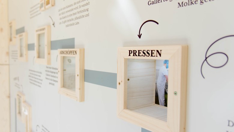 Eine wei&szlig;e Wand mit Beschriftungen und Holzrahmen, durch welche man in den Produktionsbereich einer K&auml;serei schauen kann.