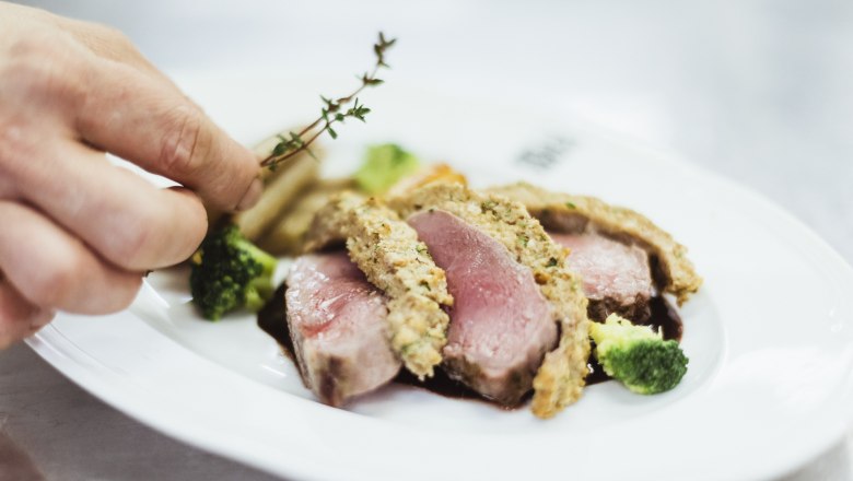 Close-up of a pink roasted meat dish with herb crust, broccoli and a hand adding thyme.