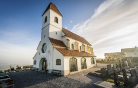 Eine wei&szlig;e Kirche mit rotem Ziegeldach und einem Friedhof im Vordergrund bei sonnigem Wetter.