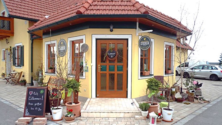 Eingang vom Bauernladen Stocker mit gelber Fassade, roten Dachziegeln und Pflanzen und Deko im Vordergrund.