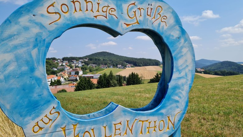 Blauer Holzrahmen in Form einer Wolke mit Schriftzug 'Sonnige Gr&uuml;&szlig;e aus Hollenthon' vor Landschaft.
