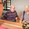 Two elderly people are sitting at a table smiling and talking in a cozy room.