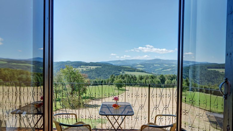 Blick von einem Balkon auf eine hügelige Landschaft mit Wäldern und Feldern, zwei Stühle und ein Tisch mit Blumen im Vordergrund.