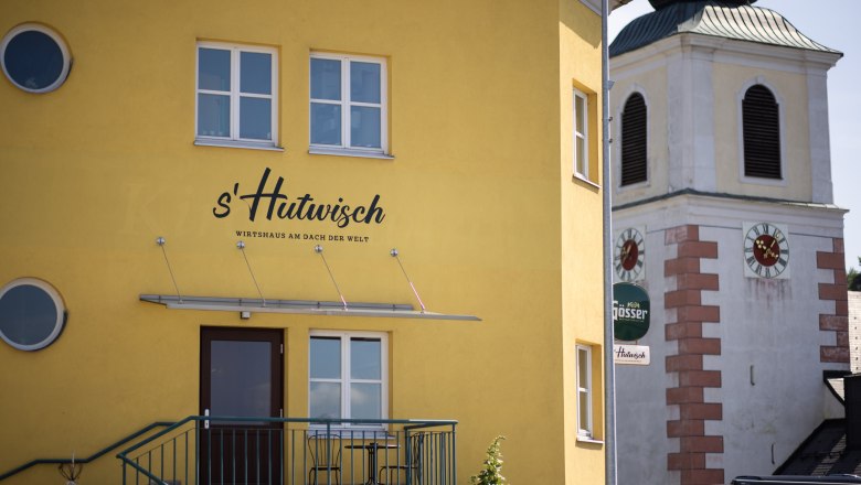 Gelbes Gebäude mit der Aufschrift 's'Hutwisch' neben einem Kirchturm mit Uhr.
