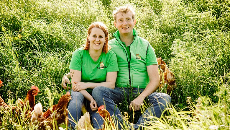 Ein Mann und eine Frau in grünen T-Shirts sitzen lächelnd auf einer Wiese umgeben von Hühnern.