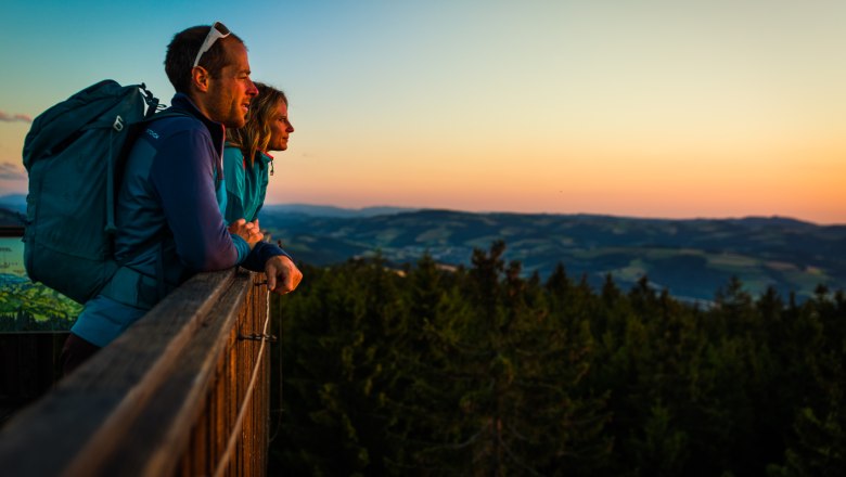 Zwei Personen stehen auf einem Aussichtspunkt und blicken in die Ferne bei Sonnenuntergang.