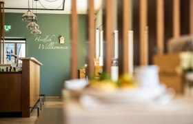 Innenansicht eines modernen Gastraumes mit grüner Wand und "Herzlich Willkommen"-Schriftzug. Über der Schank aus Holz hängen Lampen und an der Wand ein kleines Vogelhaus zur Verzierung.