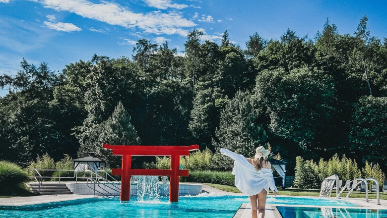 Frau in weißem Kleid und Hut geht auf einem Steg zu einem roten Torii am Pool, umgeben von Bäumen.