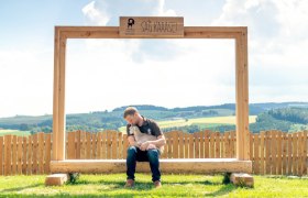 Ein Mann sitzt auf einer Holzbank unter einem großen Rahmen mit der Aufschrift 'SAG KÄÄÄSE' und hält ein Zicklein. Im Hintergrund ist eine ländliche Landschaft zu sehen.