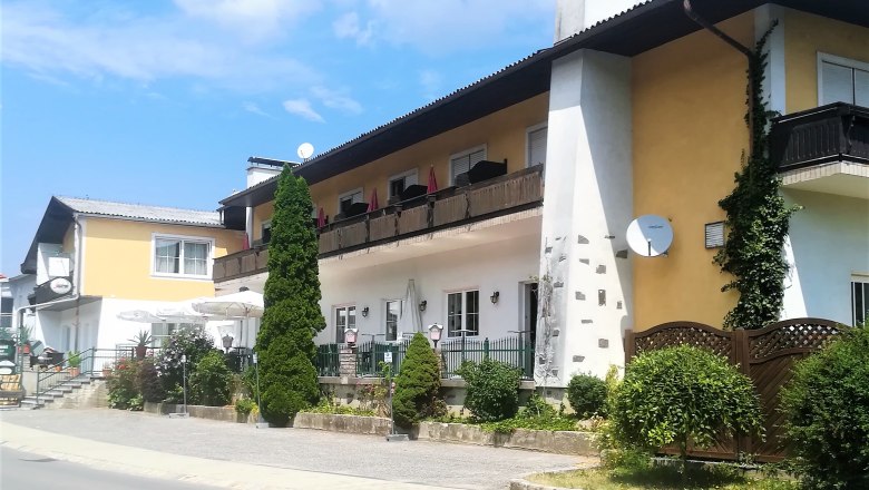 A two-storey building with balcony and terrace, surrounded by plants and trees, in sunny weather.
