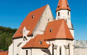 Wehrkirche Edlitz mit vielen Seitenbereichen, roten Ziegeldächern, einem spitzen Turm und blauem Himmel.