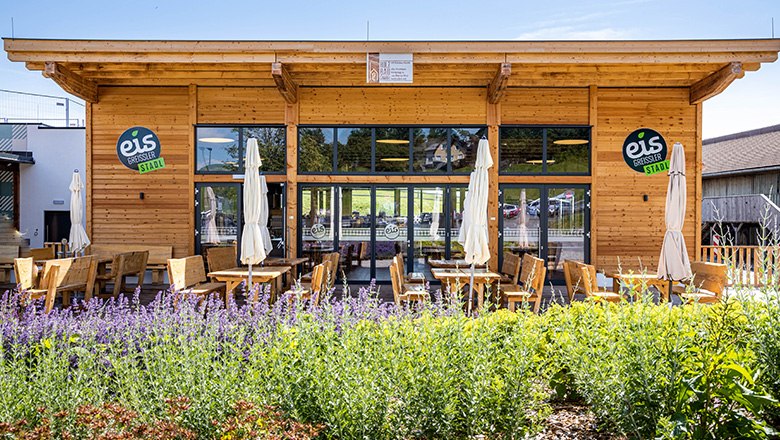 Modernes Holzgeb&auml;ude mit Terasse, auf der B&auml;nke, St&uuml;hle und Sonnenschirme stehen.