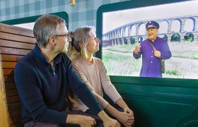 A couple is sitting in front of a screen showing a man in uniform in front of a viaduct.