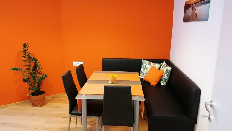 A modern breakfast room with an orange wall, black corner bench, table and chairs. A plant stands in the corner, a picture hangs on the wall.