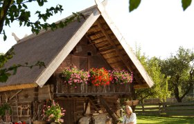 Traditional wooden building with thatched roof and flowers, woman and child in traditional costume in front.