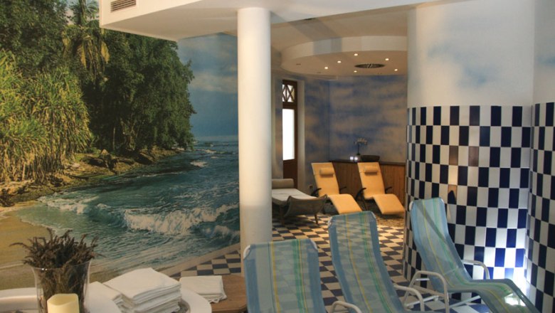 Ein Saunabereich mit Liegestühlen, einem Wandbild von einem Strand und blau-weiß karierten Fliesen.