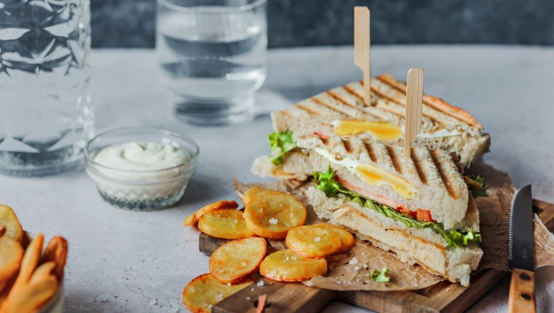 Ein Clubsandwich mit Pommes und Dip auf einem Holzbrett, daneben ein Glas Wasser.
