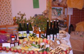 Ein Tisch in einem Hofladen mit verschiedenen Bio-Produkten wie Äpfeln, Marmeladen, Säften und Likören, dekoriert mit Blumen und Pflanzen.