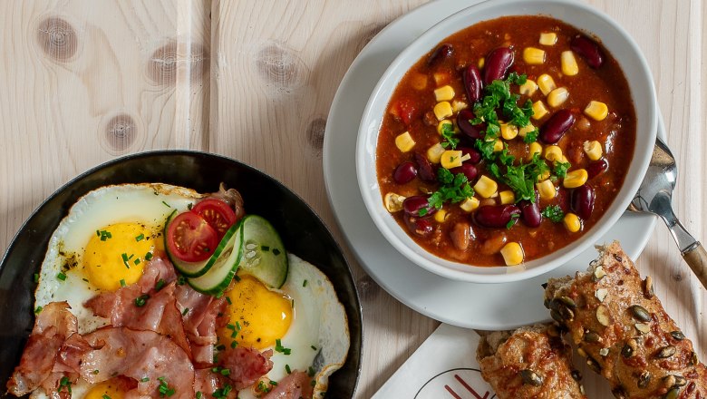 Verschiedene warme Snacks auf einem Tisch, darunter Sandwiches, Spiegelei mit Speck, Suppe und Brötchen.