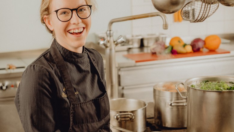 A chef in a black uniform stands smiling in a professional kitchen, frying a piece of meat in a pan.