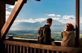 Zwei Personen genießen die Aussicht von einem Aussichtspunkt auf eine weite Landschaft mit Bergen im Hintergrund.