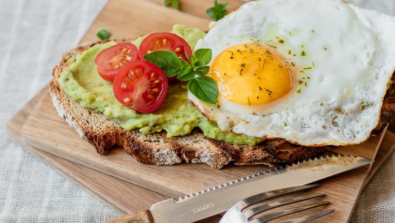 Ein belegtes Brot mit Avocado, Tomaten und Spiegelei auf einem Holzbrett.