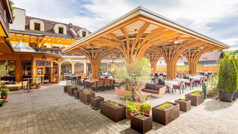 Terrasse mit Holzdach und Tischen, umgeben von Pflanzen.