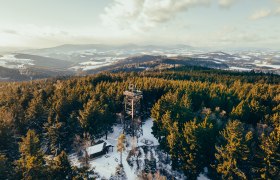 L&eacute;gi felv&eacute;tel egy h&oacute;val bor&iacute;tott tűlevelű erdőről, a h&aacute;tt&eacute;rben egy kil&aacute;t&oacute;toronnyal &eacute;s dombokkal.