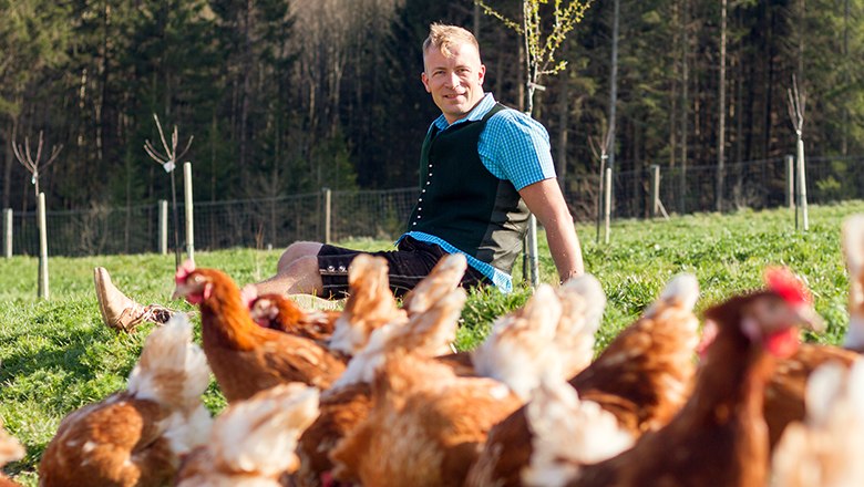 Ein Mann in Tracht sitzt auf einer grünen Wiese mit Hühnern im Vordergrund.