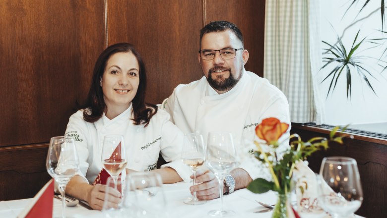 Two people in chef's jackets sit at a laid table with wine glasses.