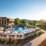 Outdoor area of a hotel with pool, parasols, sun loungers, surrounded by green meadows and trees.
