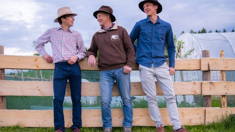 Three men in hats lean against a wooden fence in a meadow, smiling.