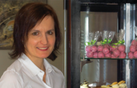 Eine Frau mit wei&szlig;er Bluse steht l&auml;chend neben einer Vitrine mit verpackter Mehlspeise.