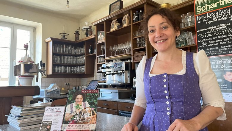 Frau in Tracht steht lächelnd hinter einer Theke, daneben ein Kochbuch und Schilder.
