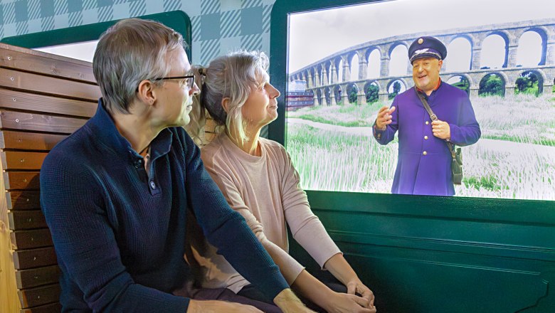 Ein Paar sitzt vor einem Bildschirm, auf dem ein Mann in Uniform vor einem Viadukt zu sehen ist.