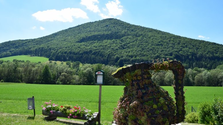 Gr&uuml;ne Landschaft mit einem bewaldeten H&uuml;gel im Hintergrund und einer bepflanzten Skulptur im Vordergrund.