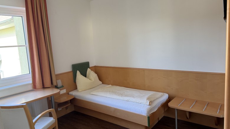 Ein einfaches Hotelzimmer mit Einzelbett, Tisch, Stuhl und Fenster mit Vorhängen.