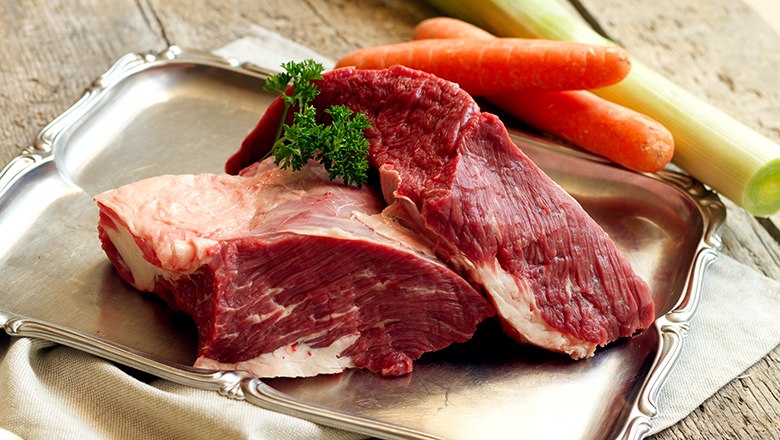 Zwei Stück rohes Fleisch auf einem silbernen Tablett auf einem Holztisch. Daneben Kartotten und Lauch.