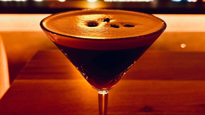 Ein Cocktailglas mit dunklem Getr&auml;nk auf einem Holztisch, im Hintergrund eine unscharfe Bar.