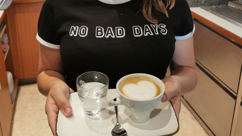 Person h&auml;lt ein Tablett mit einem Cappuccino und einem Glas Wasser.