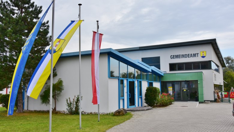 Ein modernes Gebäude mit der Aufschrift 'Gemeindeamt' und drei Fahnenmasten davor.