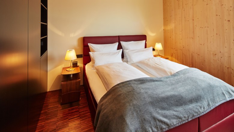 Ein gemütliches Schlafzimmer mit einem Doppelbett, roten Kopfteil, zwei Nachttischen mit Lampen und Holzwänden.